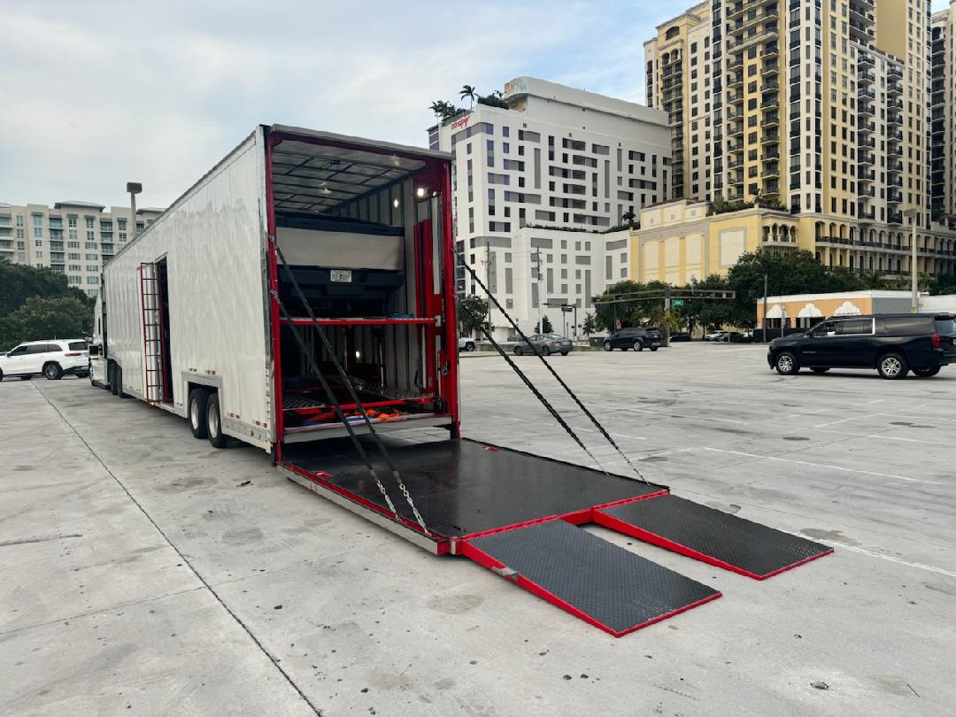 A large white enclosed multi-level car carrier trailer is parked in a paved area with its rear open, showing a vehicle on the upper deck and loading ramps extended to the ground. Tall city buildings rise in the background.