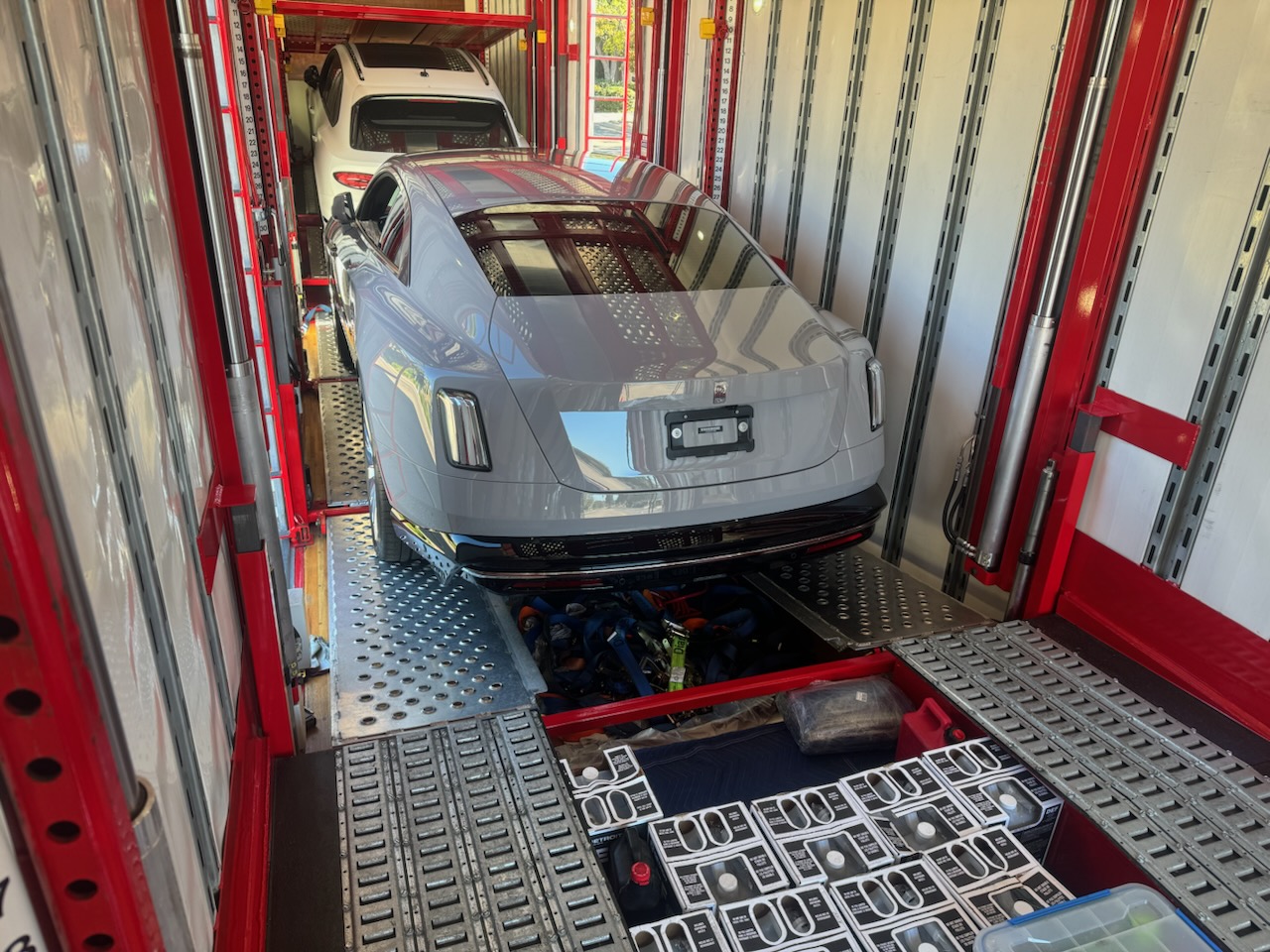 Two cars are loaded inside a red and white car transporter, with a grey luxury coupe on the lower deck and a white SUV positioned above it on the upper deck.